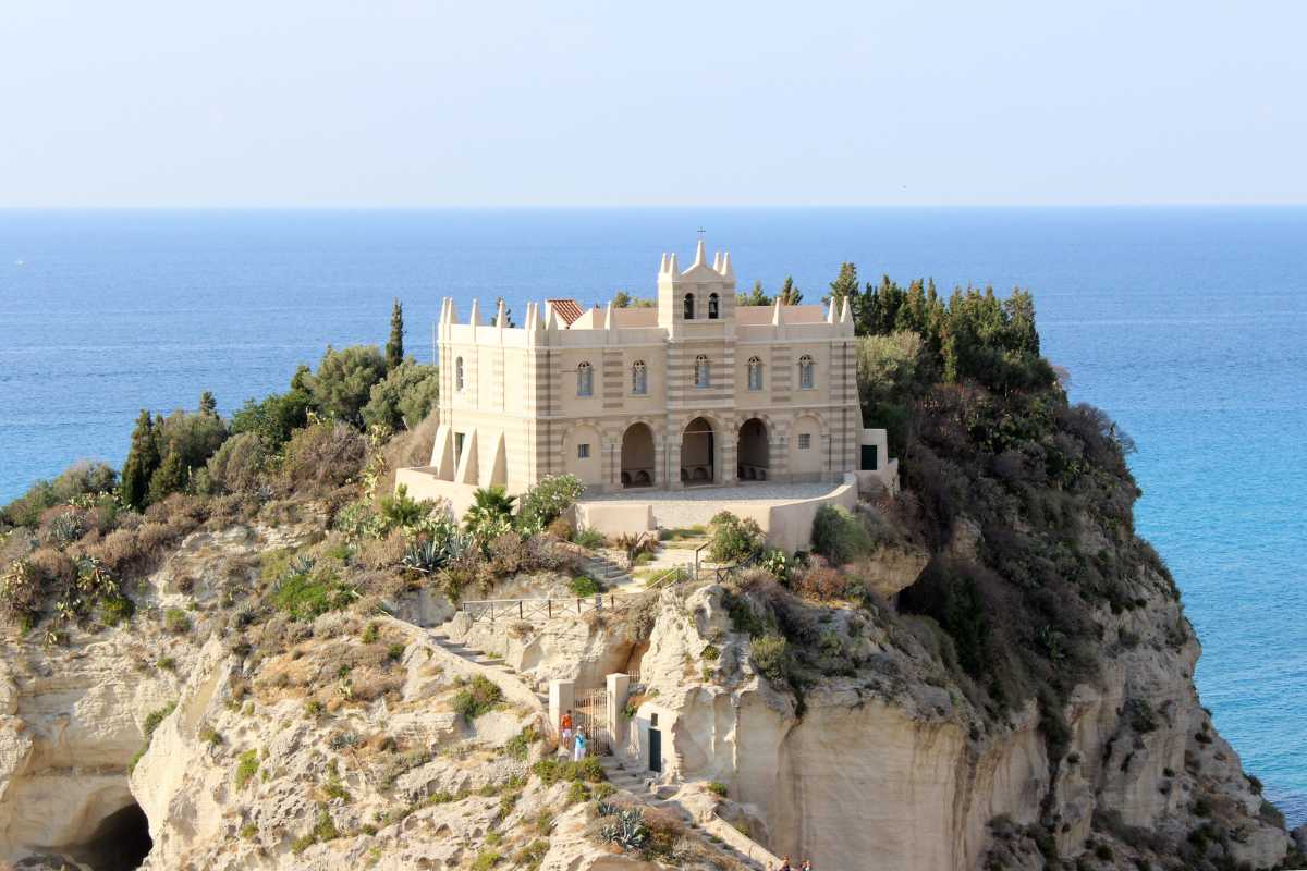 Primo piano della scalinata che porta al Santuario di Santa Maria dell’Isola con visitatori, dettagli della roccia e mare sullo sfondo, luce estiva